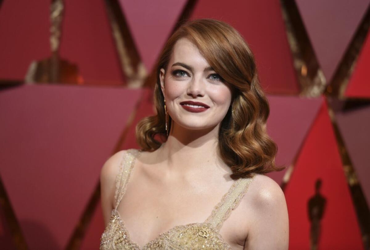 Emma Stone stands in front of a red background in a sparkly, strappy pale gold gown and wavy red hair