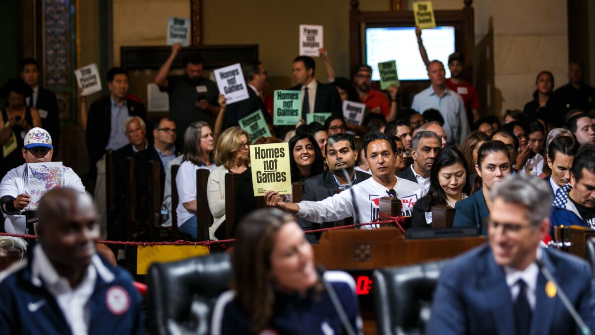 Protesters hold up signs as the representatives of the L.A. 2028 Olympic bid committee speak before the City Council on Thursday.