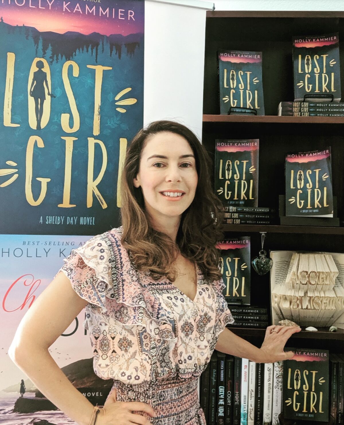 Author and publisher Holly Kammier stands in front of a poster and shelf promoting her third book, “Lost Girl, A Shelby Day Novel,” which is scheduled for release by Acorn Publishing on Jan. 5.
