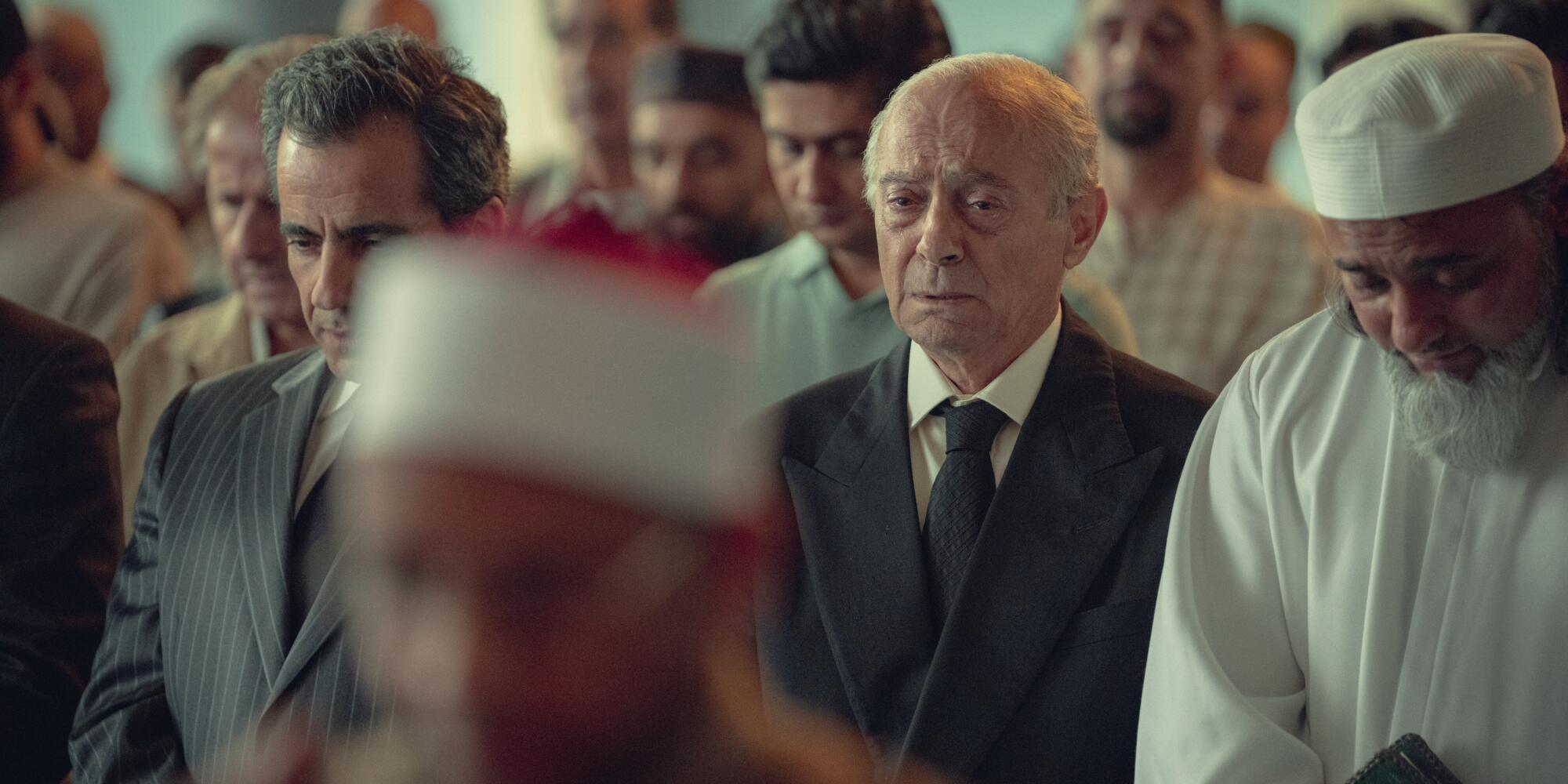 Salim Daw as Mohamed Fayed, with a solemn look on his face and tears in his eyes, stands between mourners in a mosque.