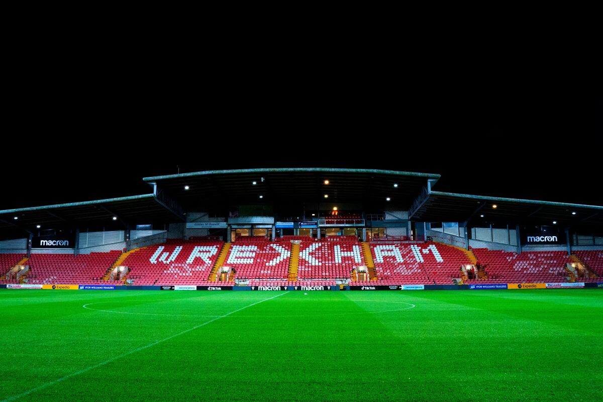 A soccer field and stadium with the word "Wrexham" in white amid the red seats.