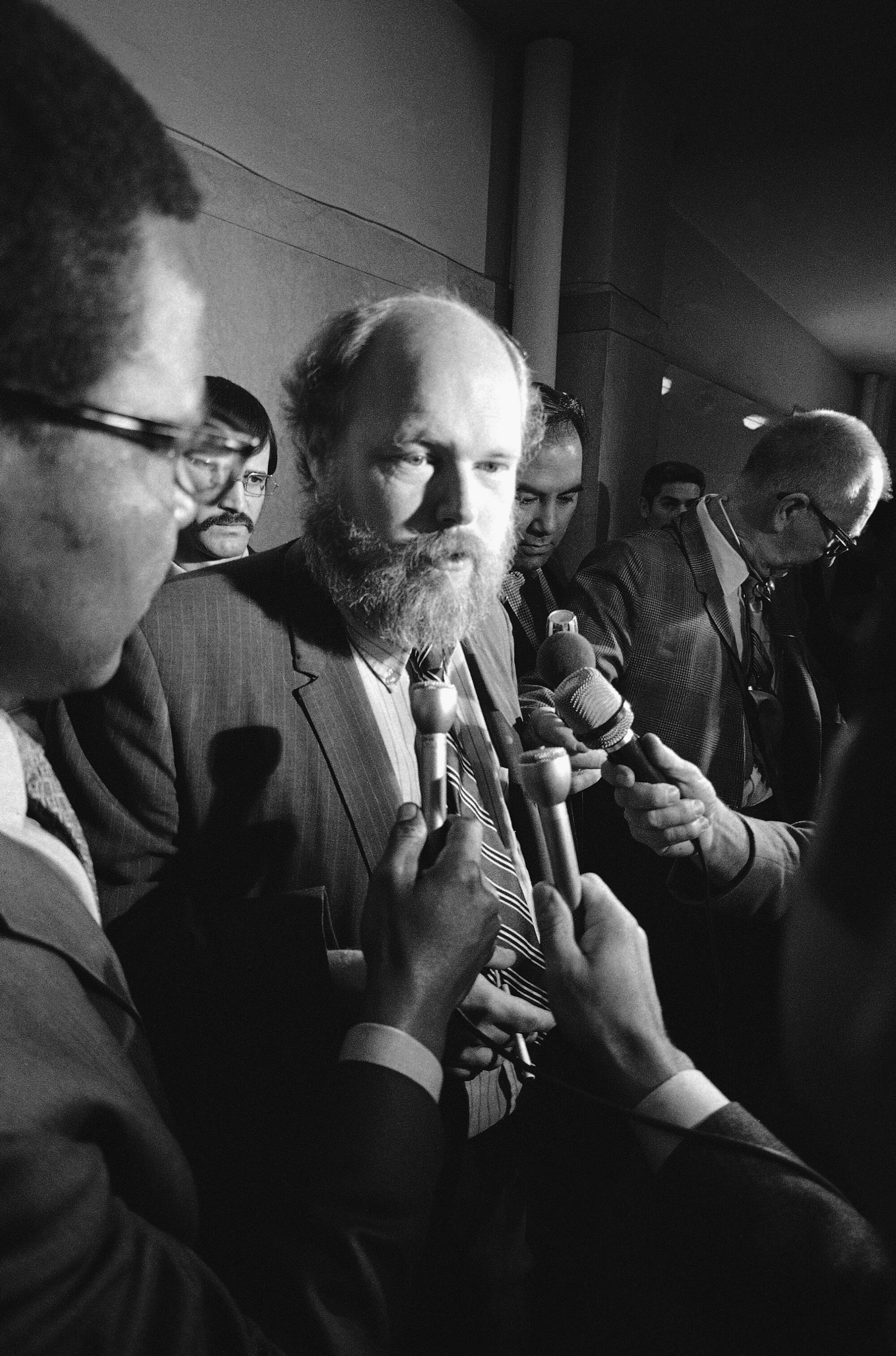 A bearded man speaks to people holding microphones.