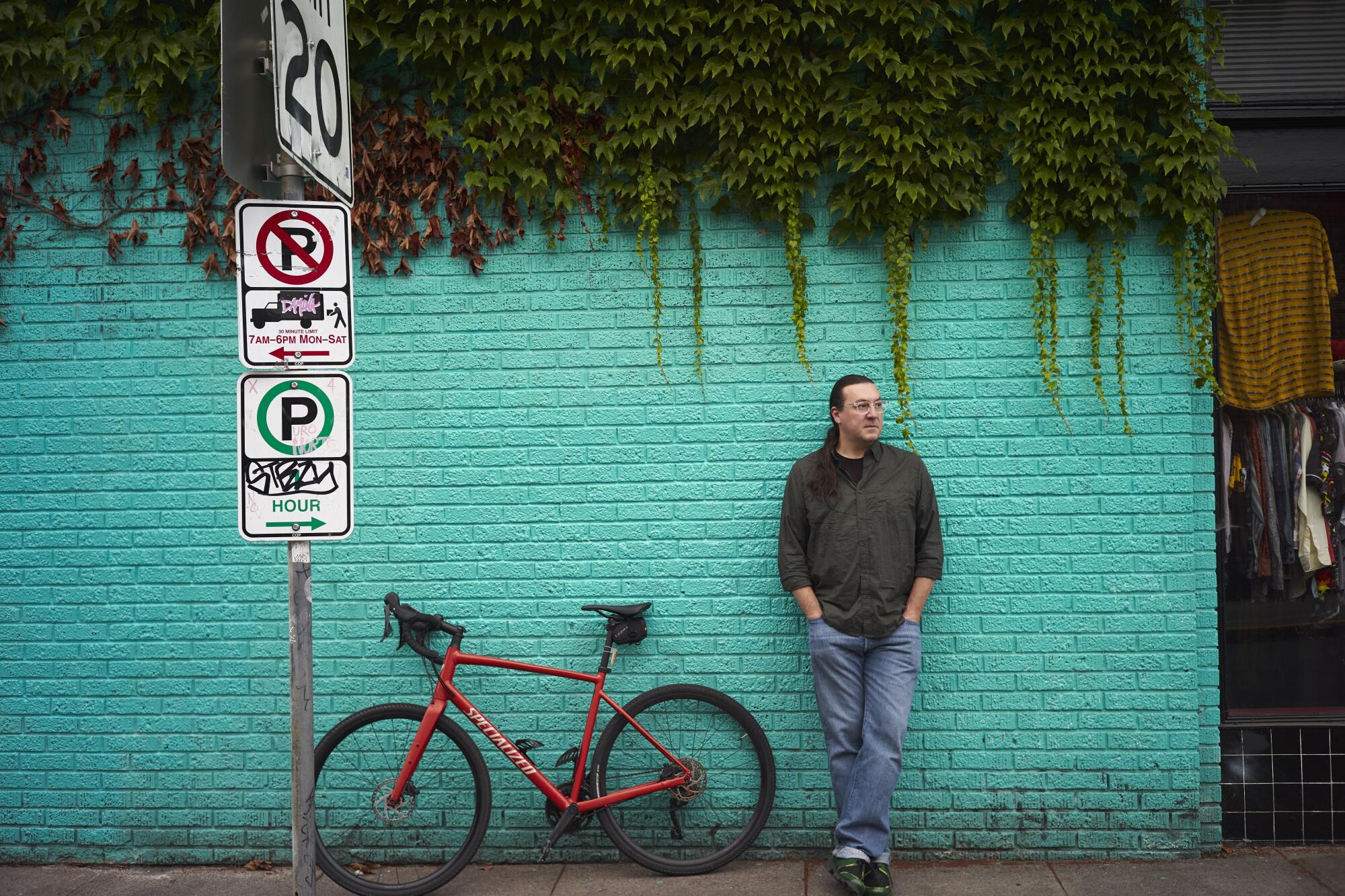 A man stands in front of a turquoise brick building with his red bike. 