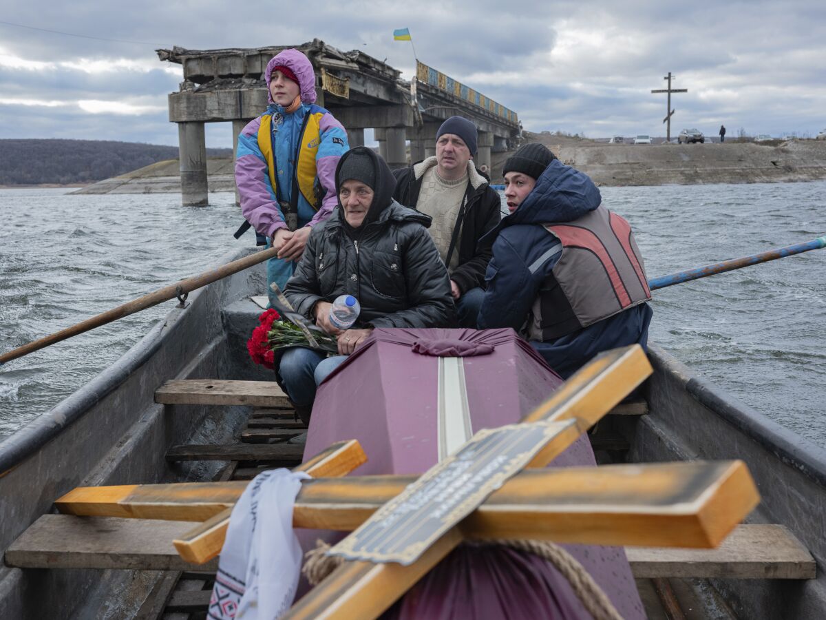 A woman sits with others and a coffin in a boat crossing a river, with a ruined bridge in the background.