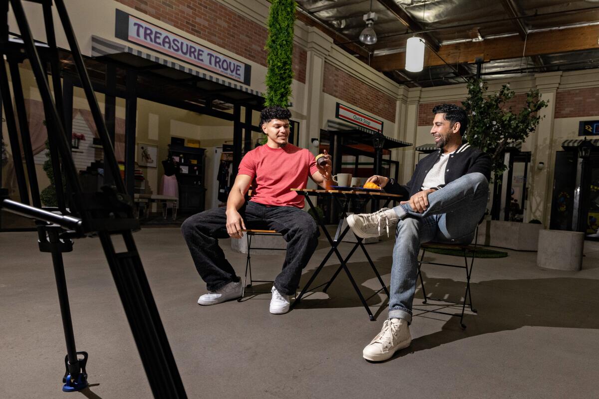 Two men, one in an orange shirt, sit facing each other across a table in a stage