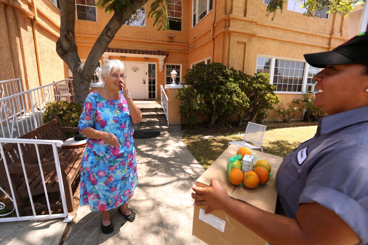 A woman stands, a hand to her mouth, outside an orange stucco building as another approaches carrying a box with fruit on top