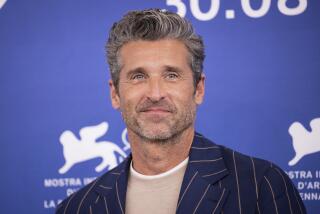 Patrick Dempsey with greying hair smiling in a tan shirt and a black, striped suit jacket against a blue backdrop