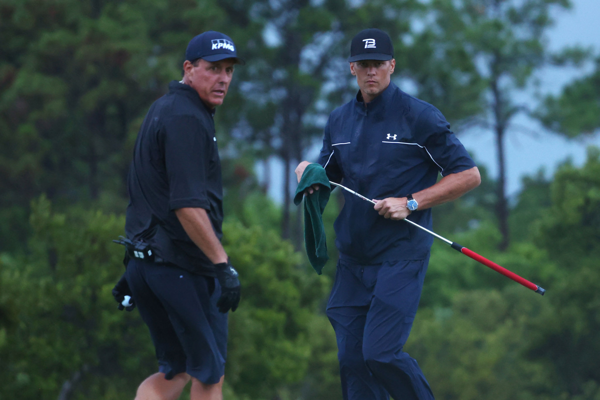 Phil Mickelson reads a putt for Tom Brady, who holds a golf club