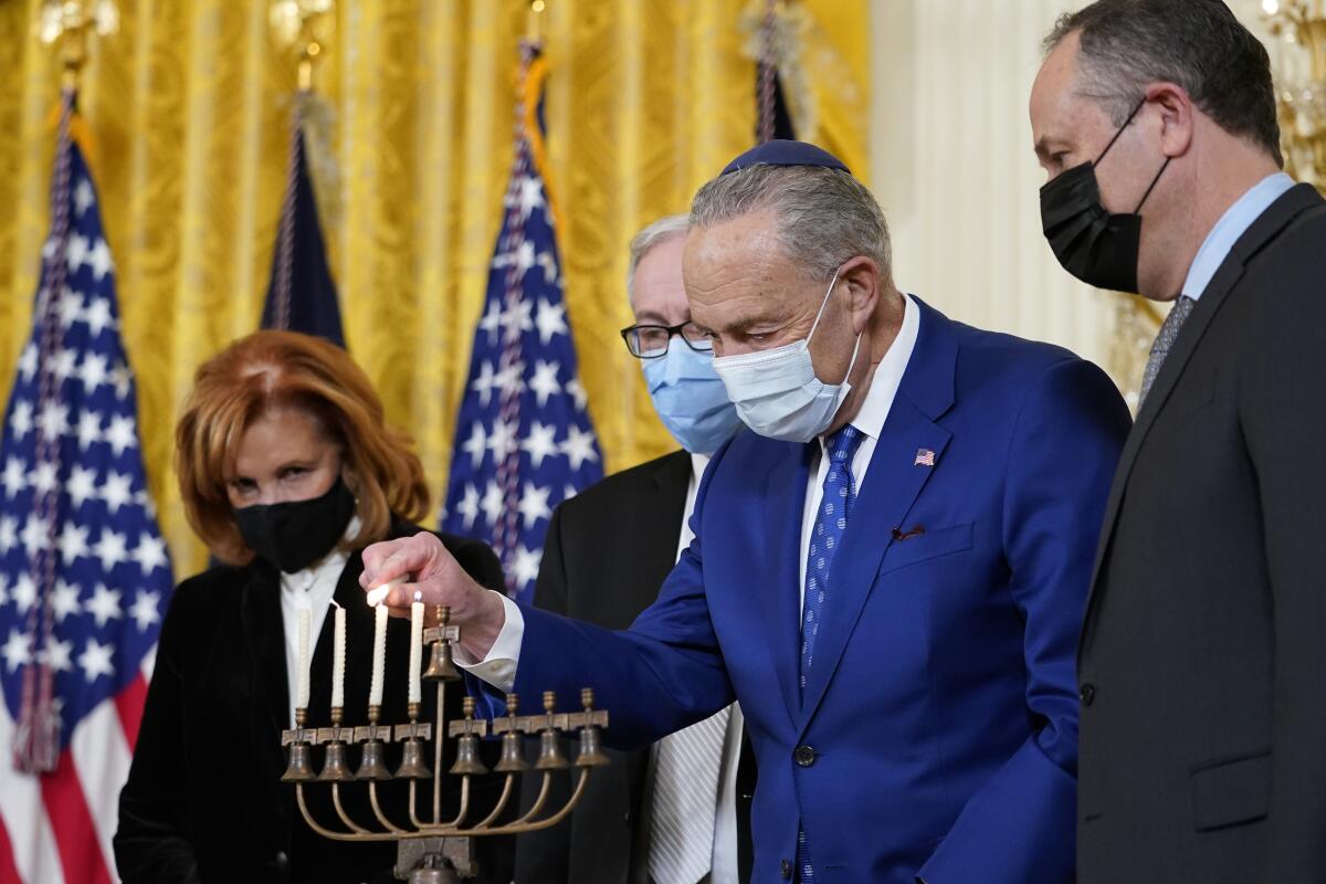 A masked man in a blue suit lights one of the four candles on a menorah as he is flanked by other people, also masked.
