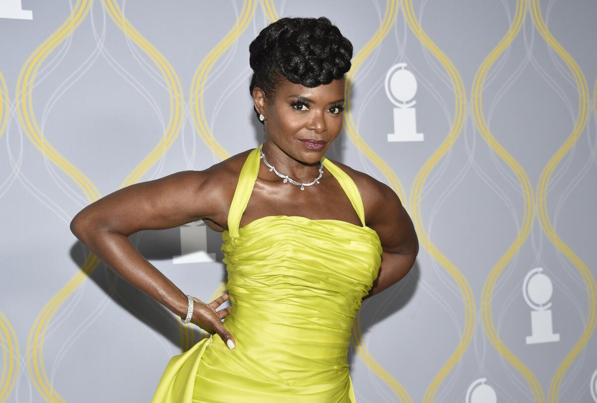 LaChanze in a bright yellow halter dress posing against a gray background with her elbows back and hands on her hips