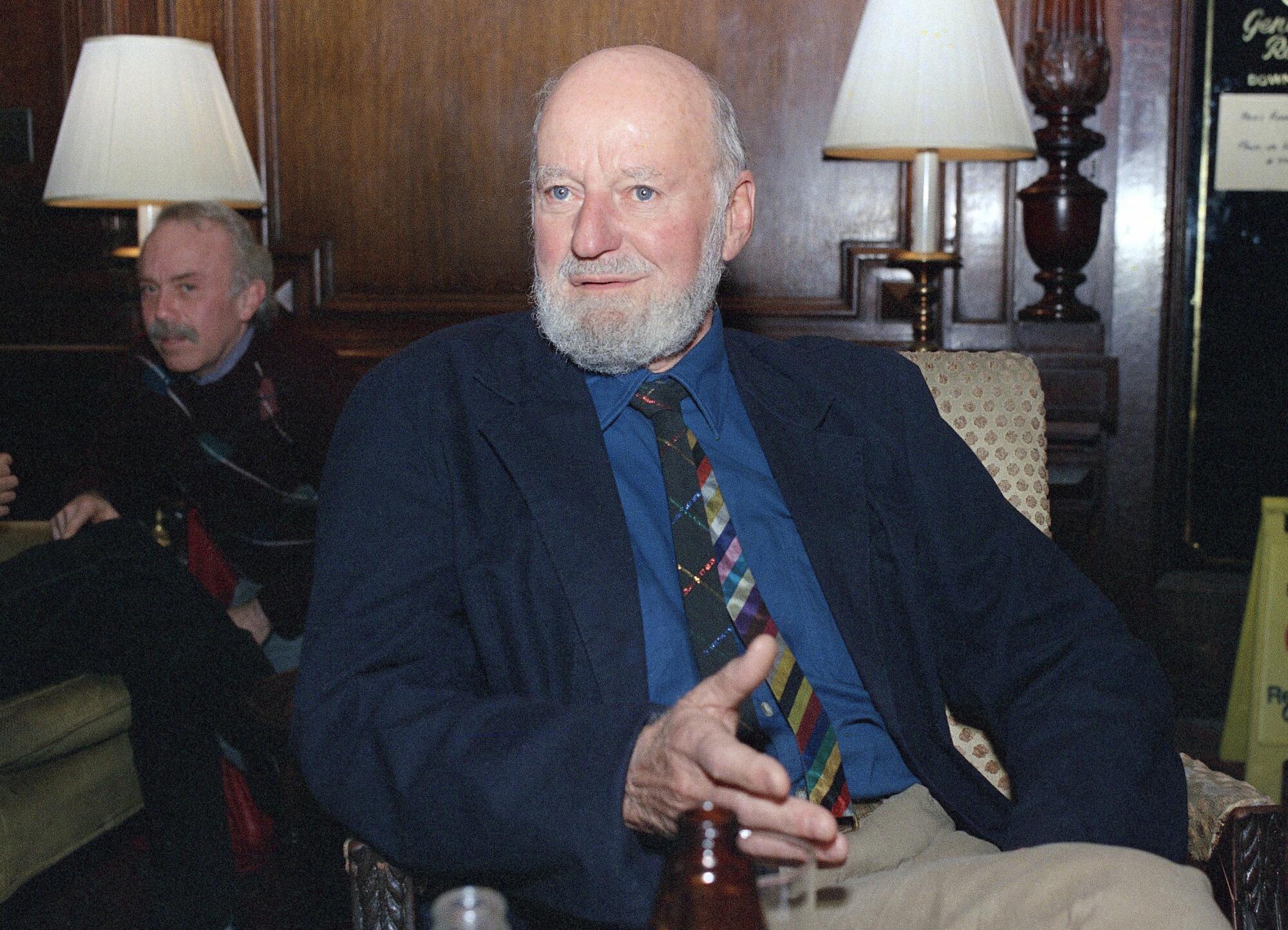 A man with a full beard in a blue jacket, striped tie and khaki pants, seated.