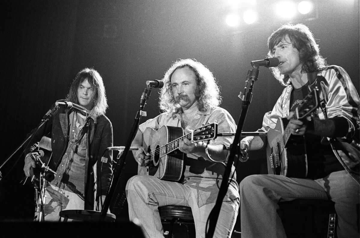 Three men with long hair on a stage. The two men on the right are sitting and holding guitars