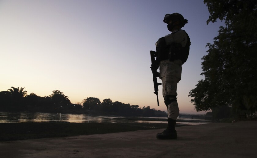 A man in military gear and holding a rifle is silhouetted at sunset alongside a river.