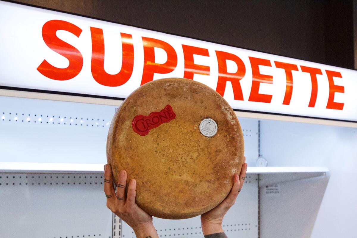 A hand holds up a wheel of cheese in front of a fridge that says SUPERETTE at DTLA Cheese Superette.