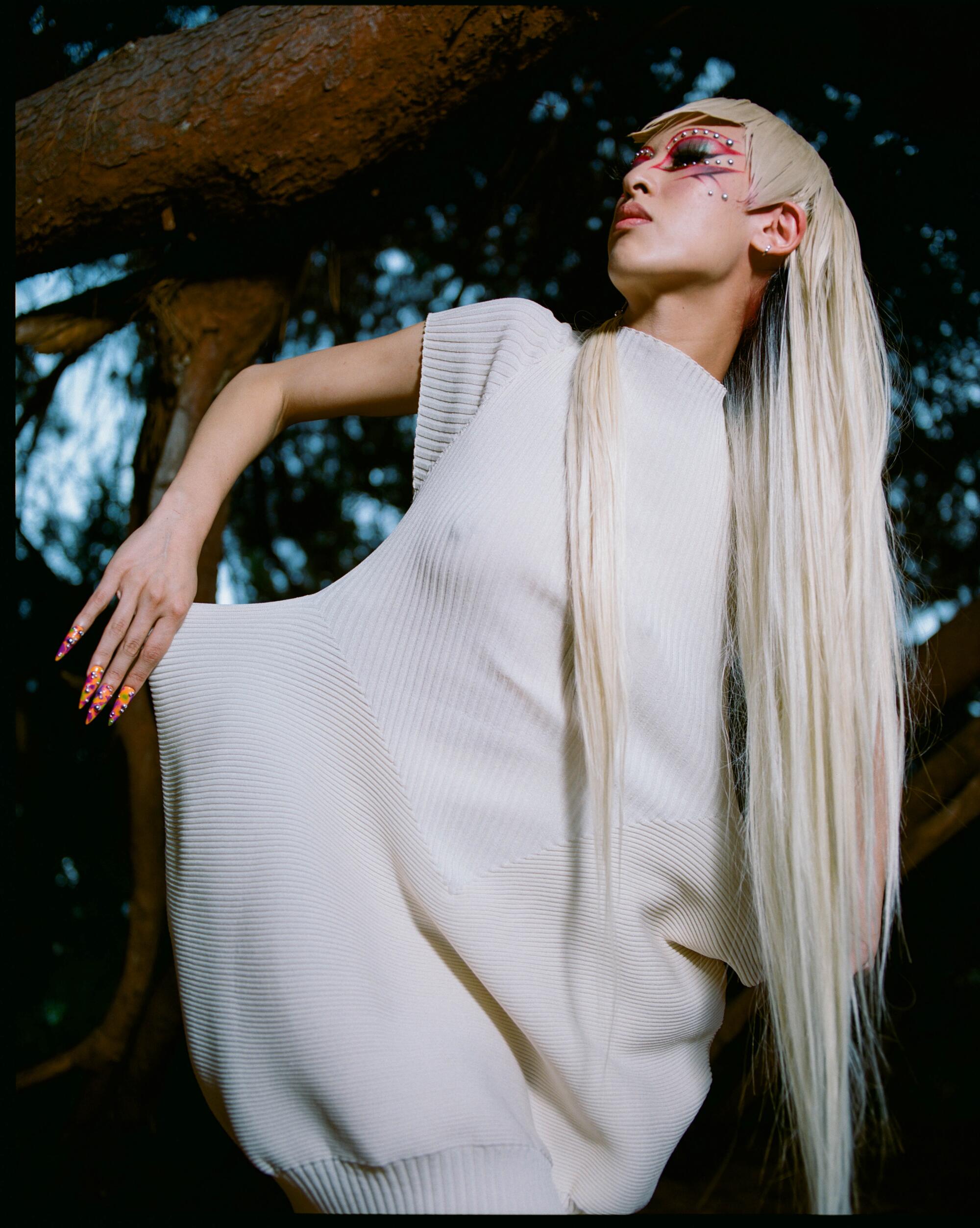 A closer view of a person with long white-blond hair standing outside in a white Issey Miyake outfit.