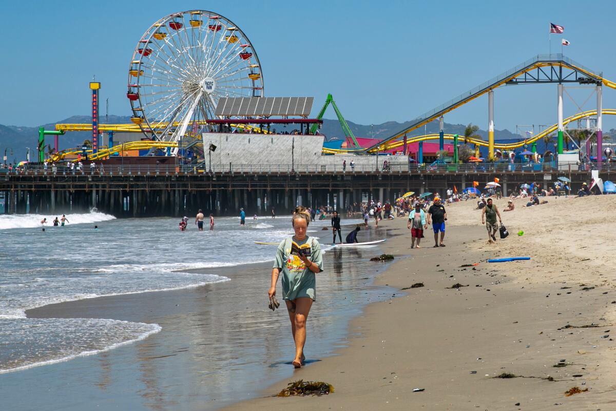 A woman reads a book as she walks near the water on a beach, people and a pier with colorful attractions behind her