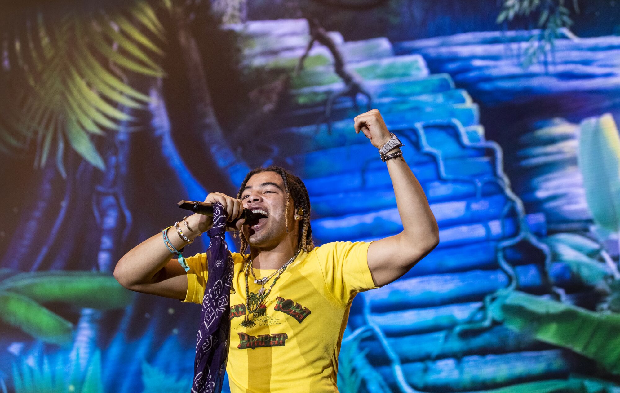 A man in a yellow shirt sings into a mic, which he holds in his right hand along with a bandana, and raises his left hand 