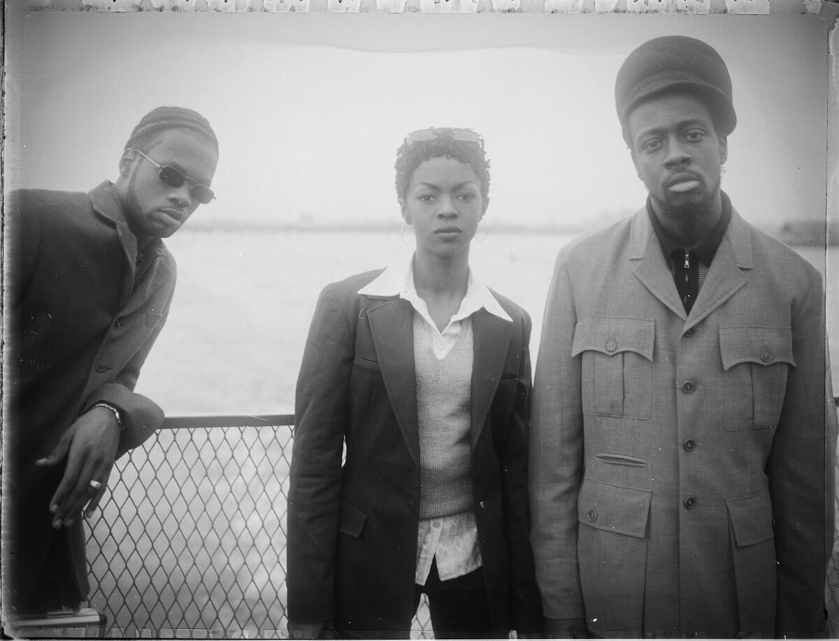Pras, Lauryn Hill and Wyclef Jean of hip-hop trio the Fugees standing in a line, posing together in front of a fence 