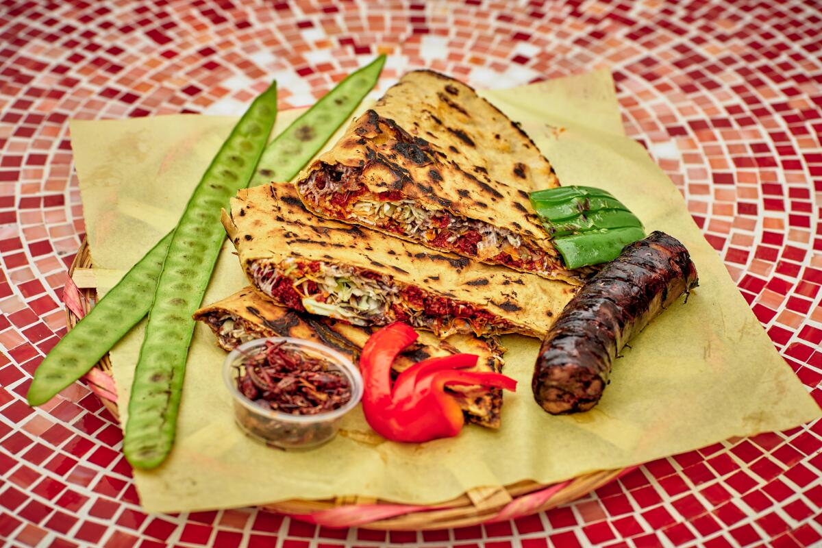 A photo of filled tortillas, two long bean-like pods, a red garnish and a sausage link on a plate, on a red mosaic table