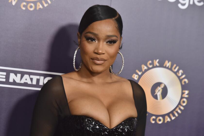 Keke Palmer with a slicked-back hairdo in a black gown and large hoop earrings posing against a dark background