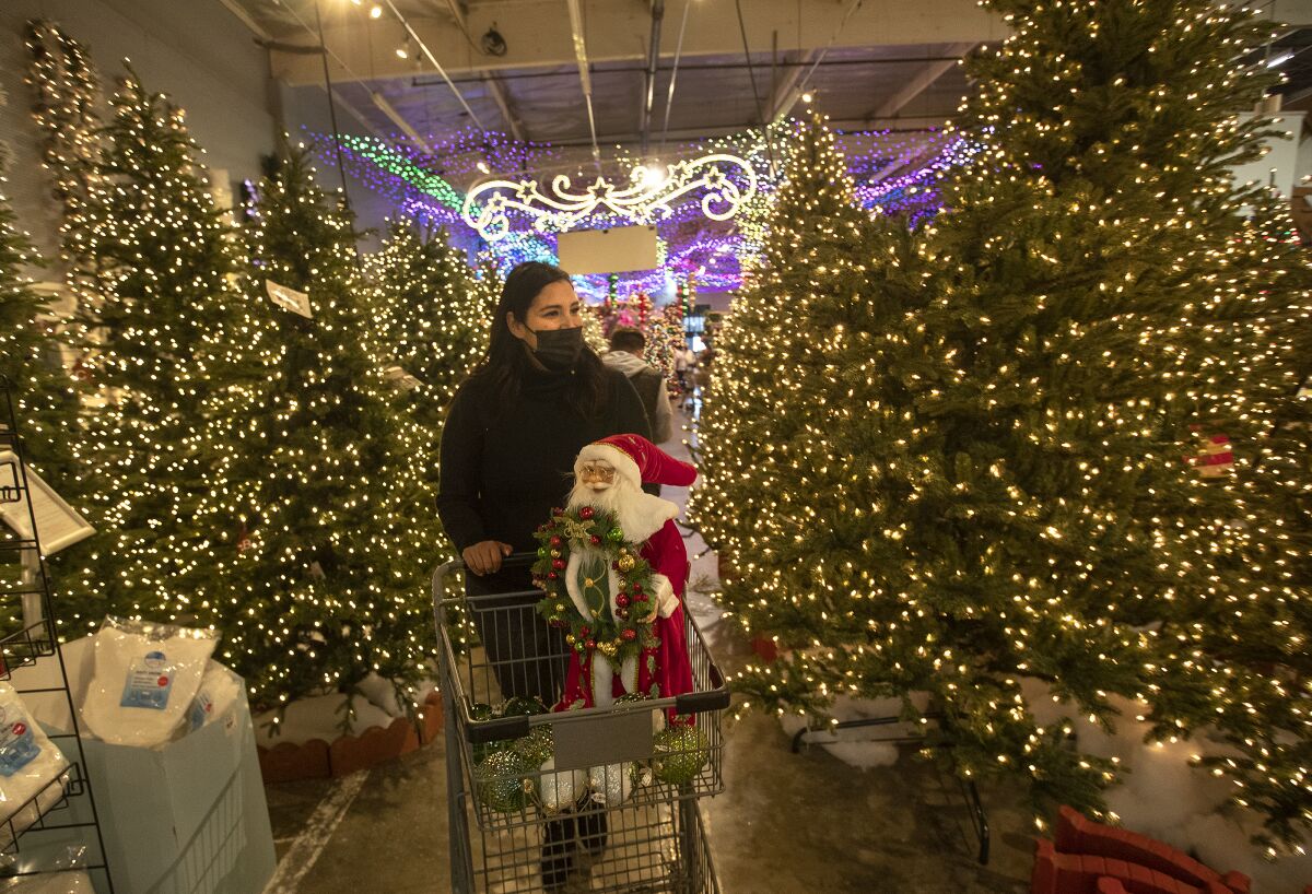 A woman pushes a shopping cart containing a Santa figurine past a row of illuminated Christmas trees
