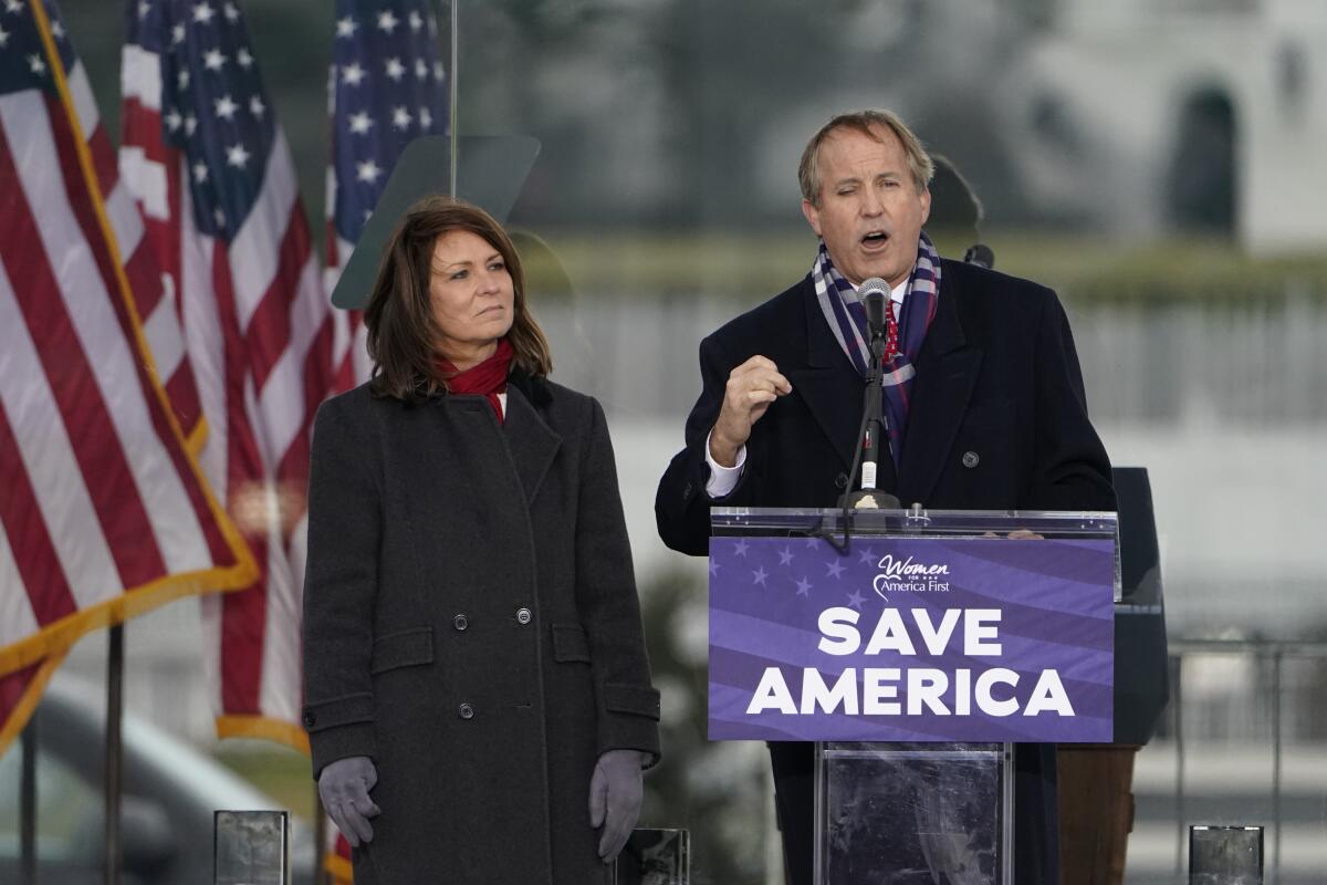 A man stands at a lectern with a sign reading "Save America" while a woman watches him.