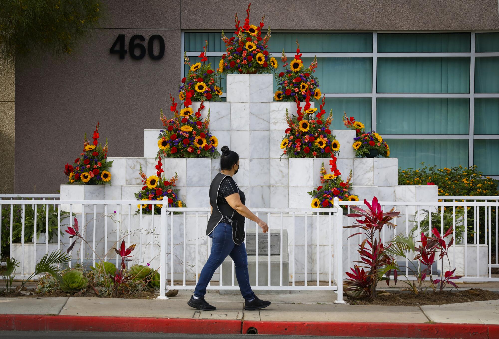 A woman walks by a memorial made up of white marble towers topped with bright floral arrangements.