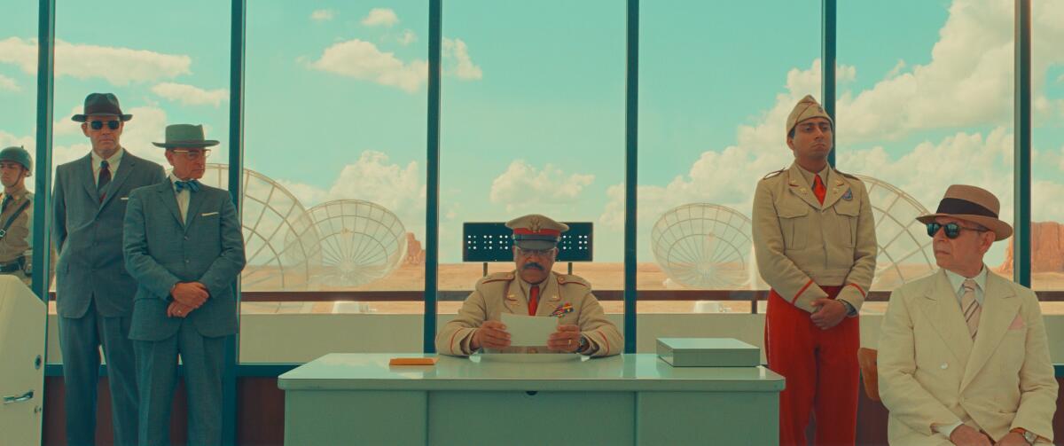 Men in various uniforms and hats sit in front of a glass wall that looks onto a field of satellite dishes.