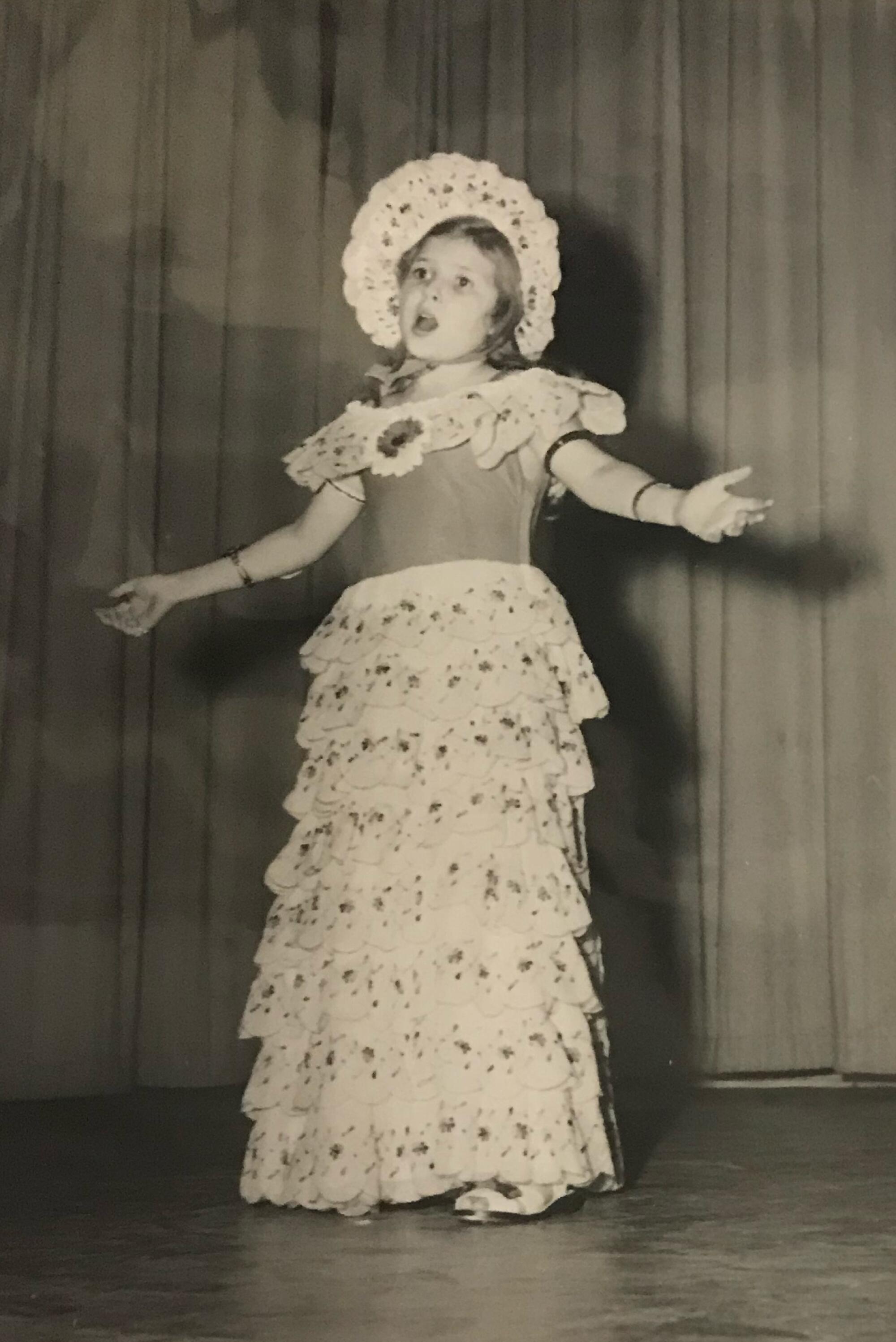 A girl in a dress and bonnet performs on a stage.