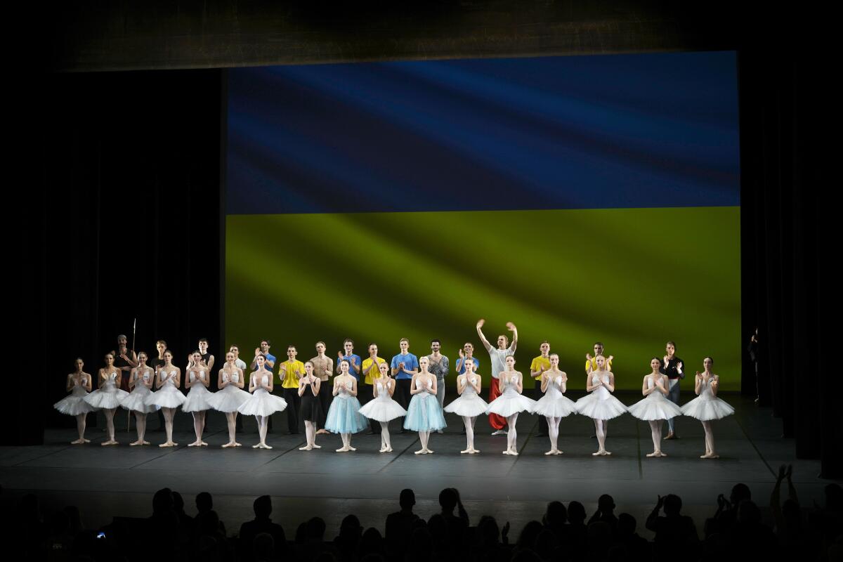 A line of ballet dancers stand onstage in front of Ukraine's flag