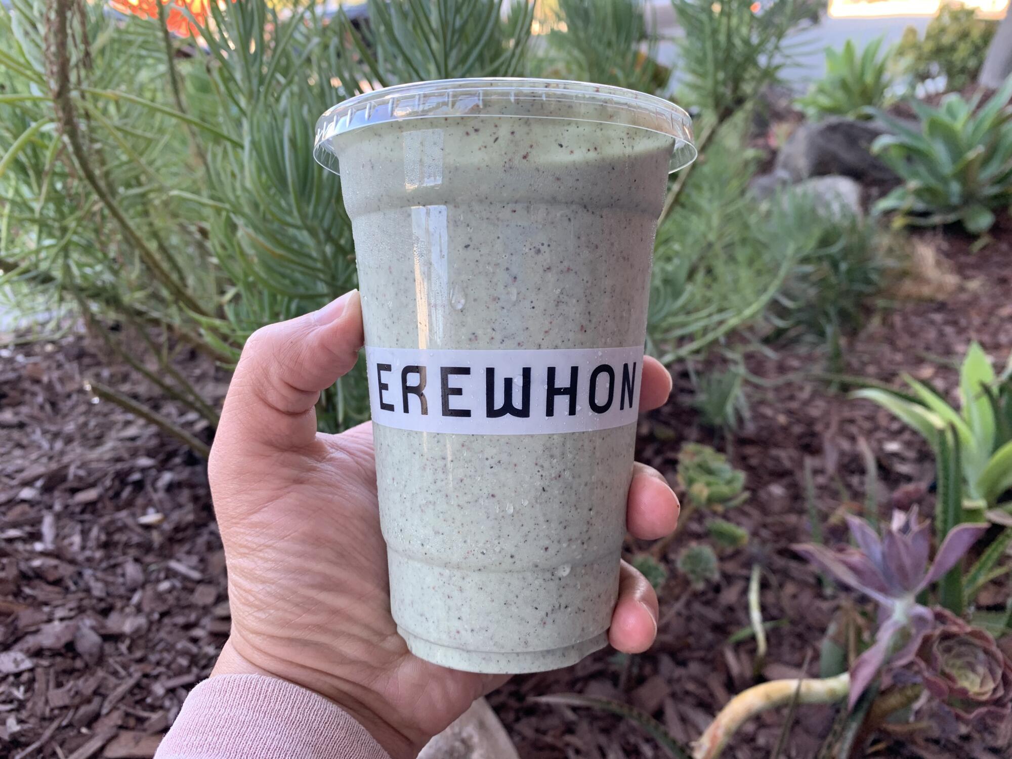 A hand holds a grayish smoothie with black specks in a plastic cup that says Erewhon.