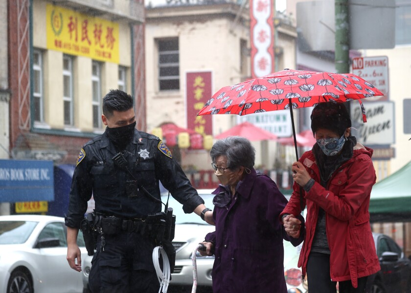 A police officer in a mask holds the arm of an elderly woman being helped on her other side by another woman with an umbrella