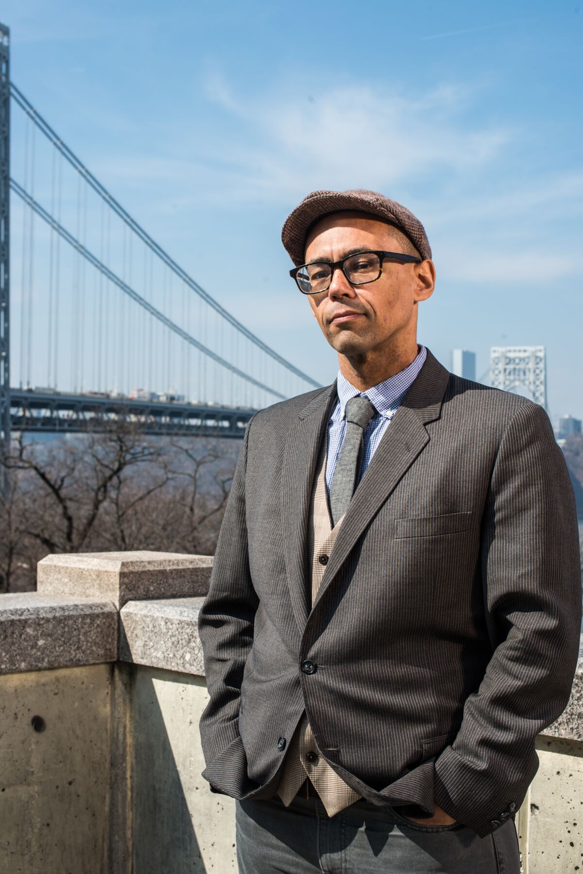 Victor LaValle wearing a suit and newsboy cap in front of a suspension bridge