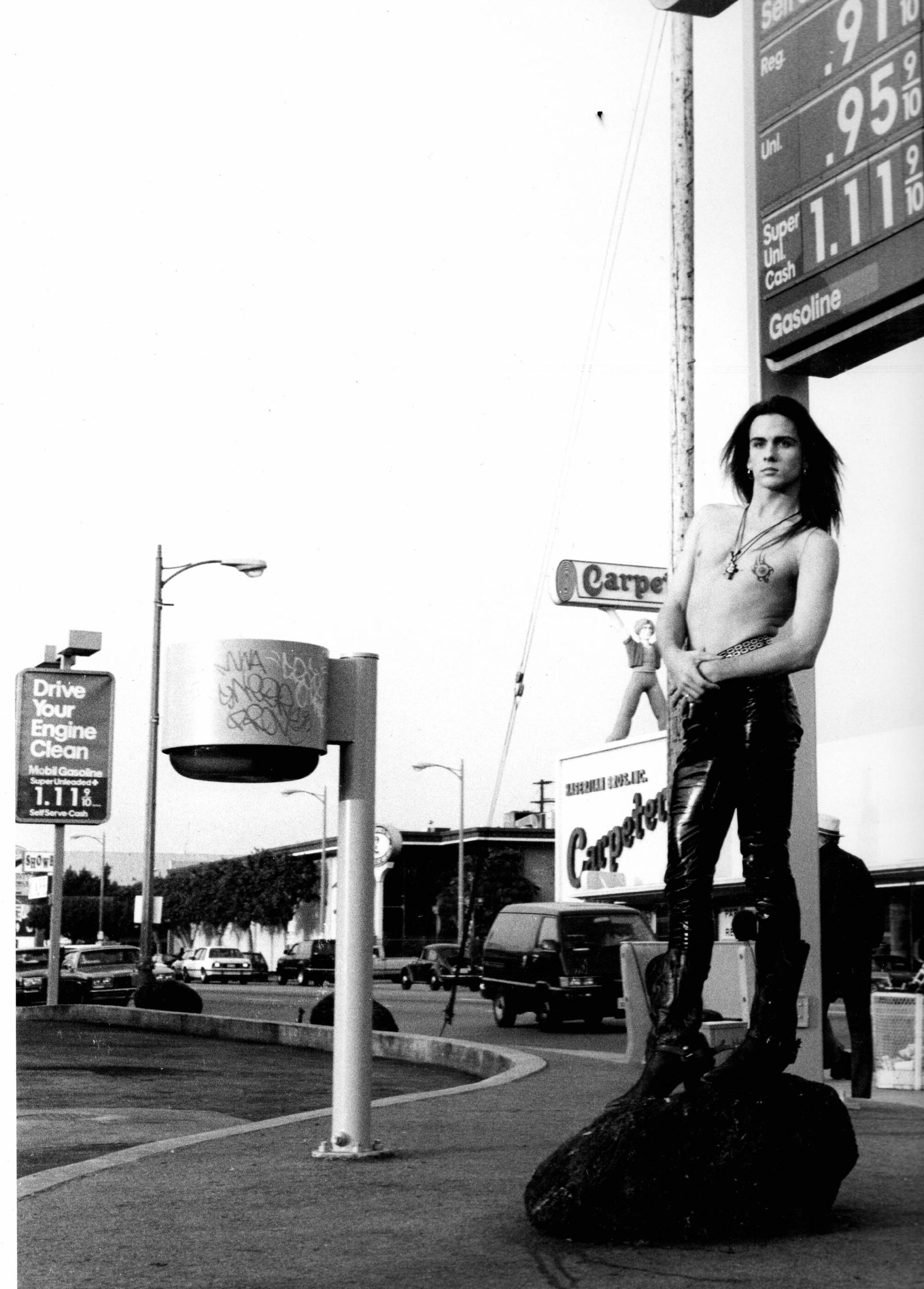 A black and white photo of a shirtless man in vinyl pants standing on a corner in front of a Los Angeles gas station.