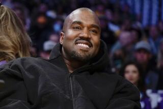 Kanye West leans back as he smiles in a black hoodie while sitting in a crowd