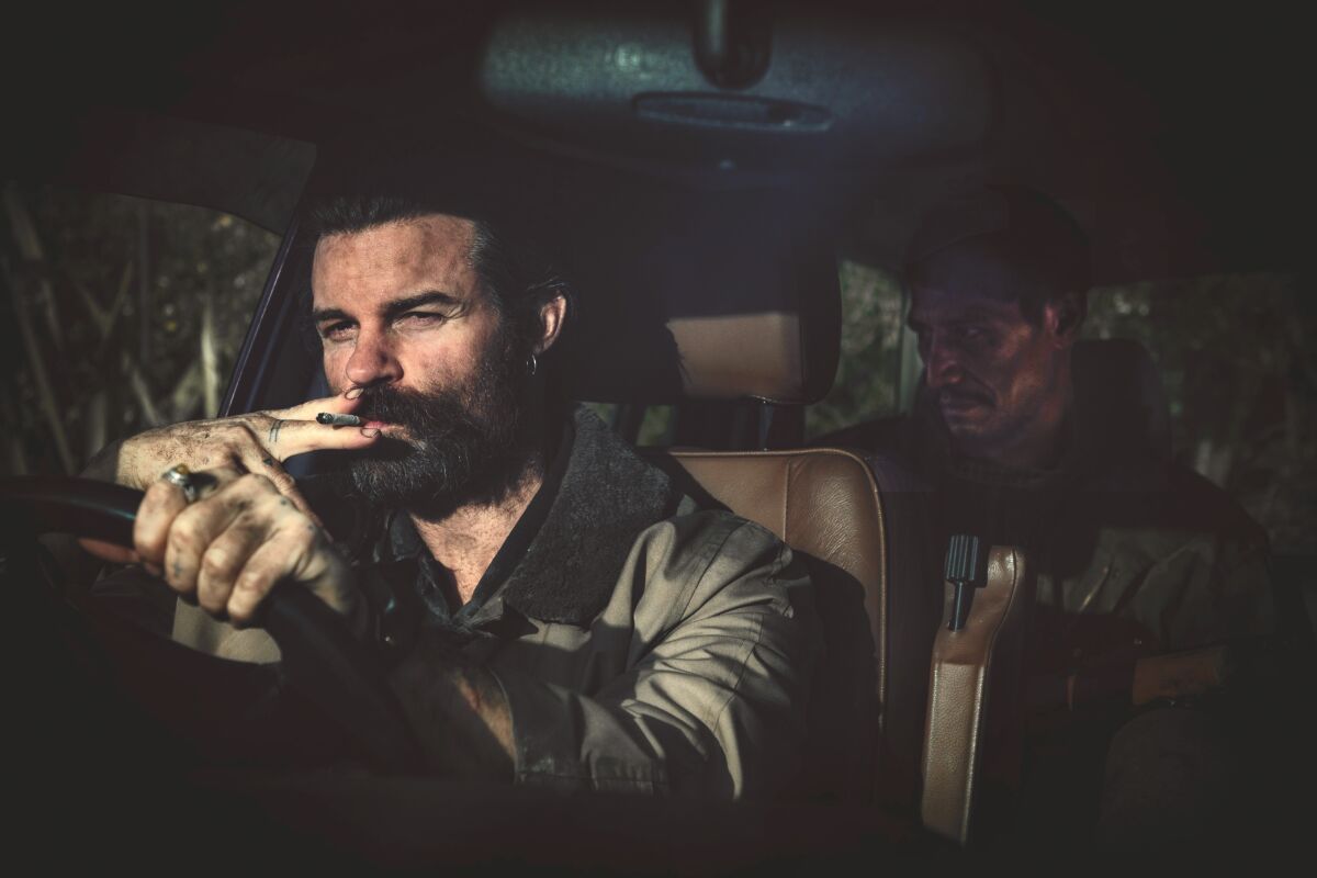 A man smokes while driving, with another man in the back seat in the movie “Coming Home in the Dark.”