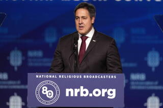 A man in a dark suit and red tie speaks at a lectern
