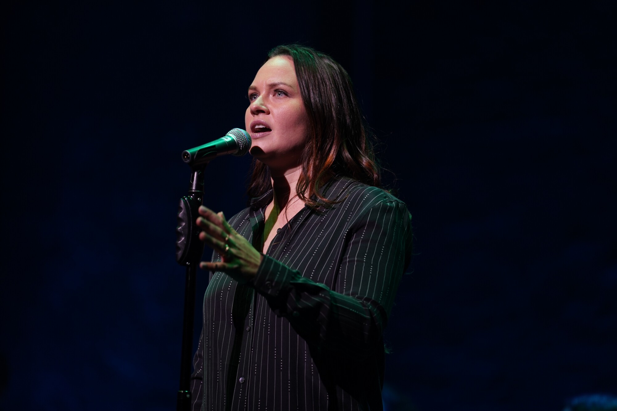 A woman wearing black sings into a microphone on a dark stage.