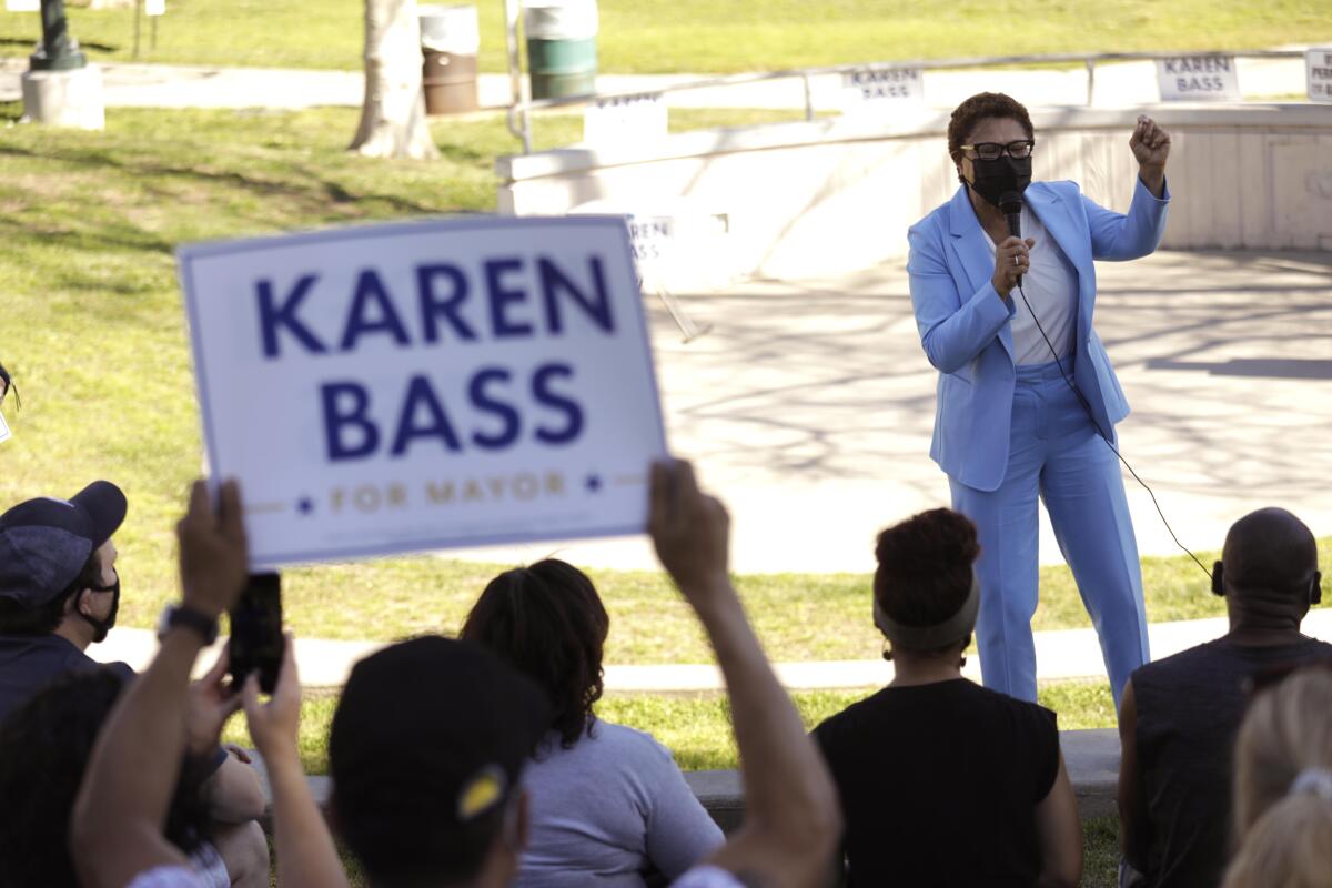 A woman in a pant suit stands outdoors and speaks to a crowd, one of whom holds a "Karen Bass for Mayor" sign.