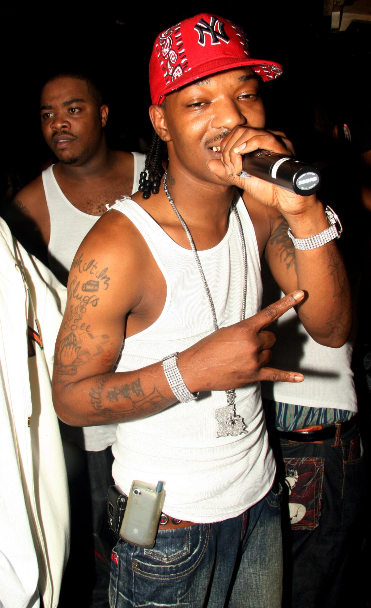 Rapper BG, in a white tank top and red Yankees cap, smiles while holding a mic to his mouth