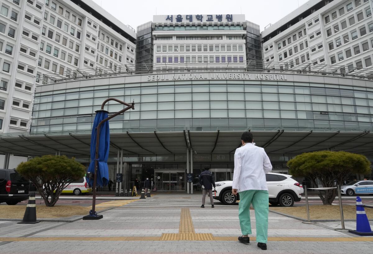 A medical worker in scrubs and a white coat walks toward a multistory building 