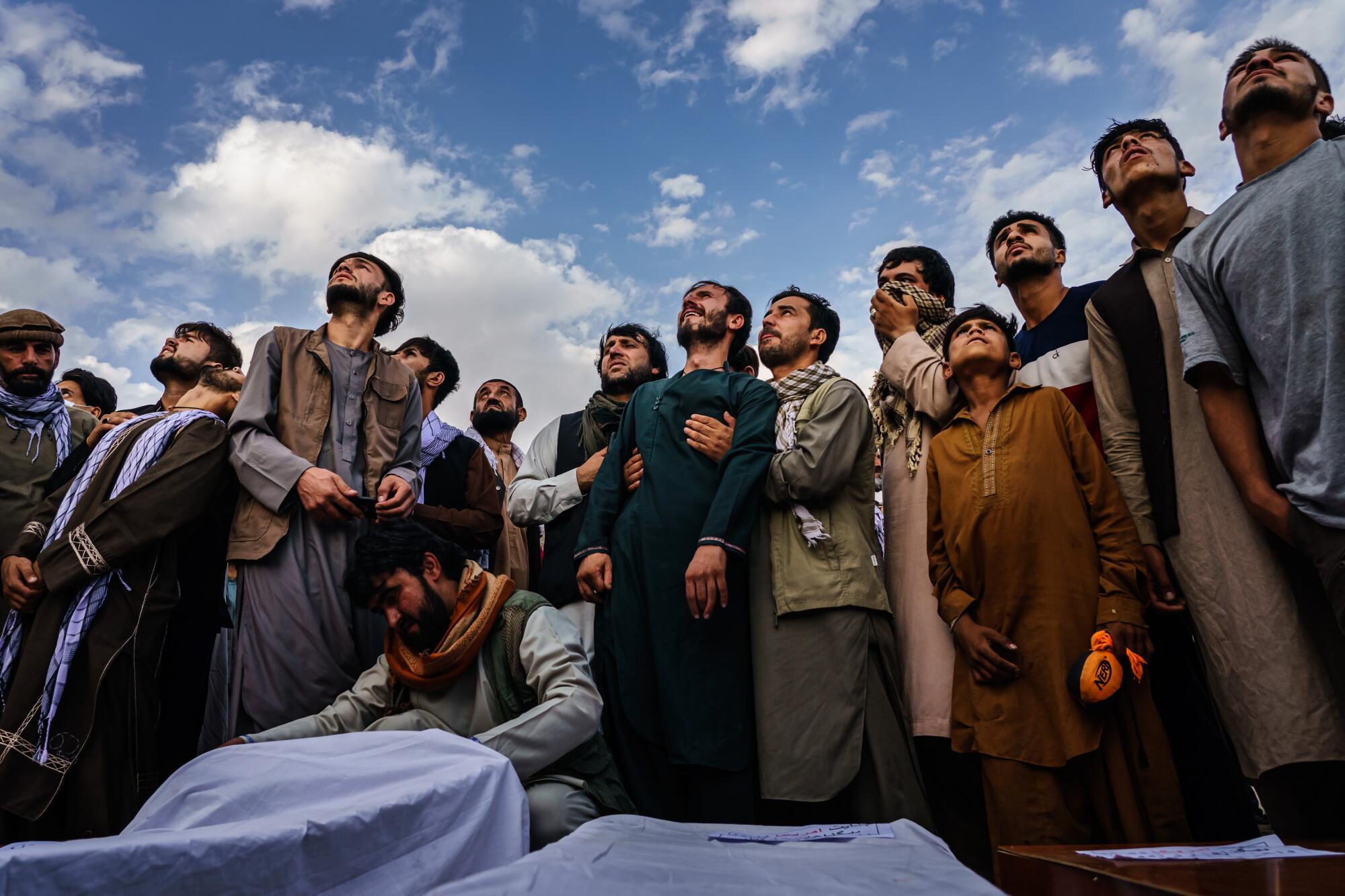 Men in tunics and pants in a winding line on a hillside attend a mass funeral