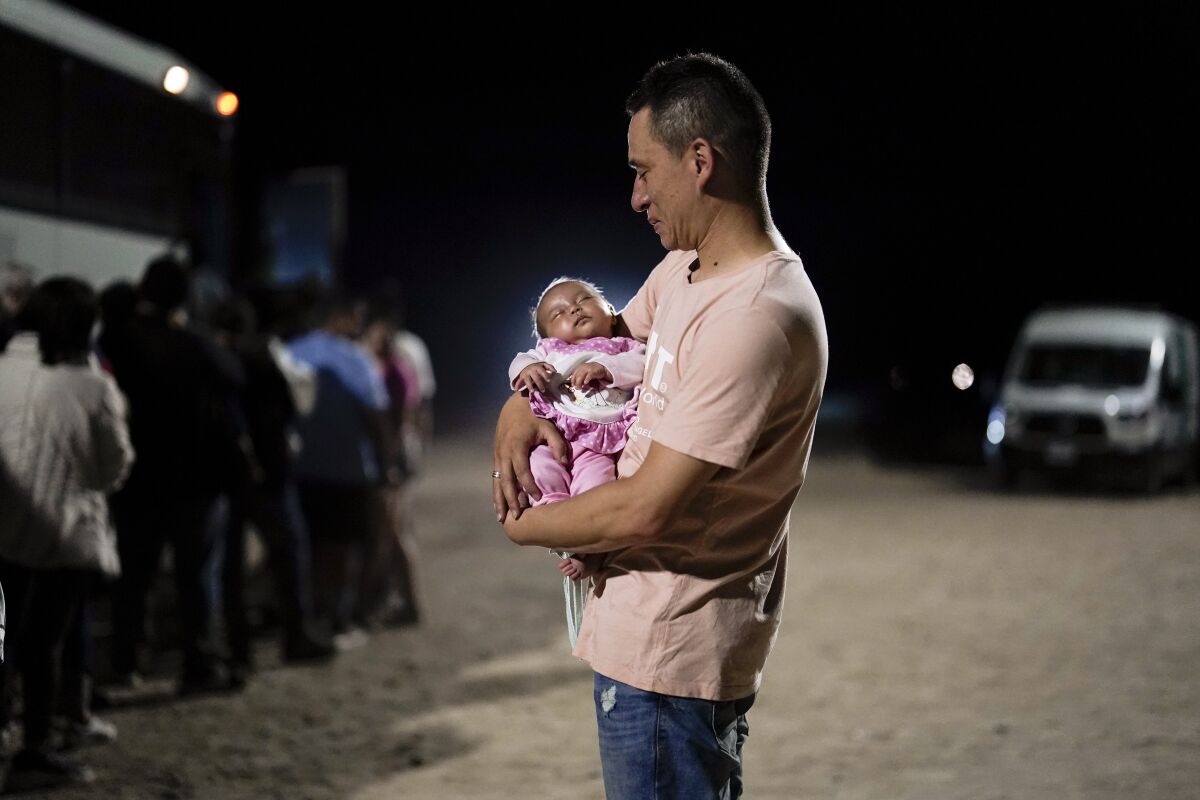 A man holds a baby while people line up next to a bus nearby