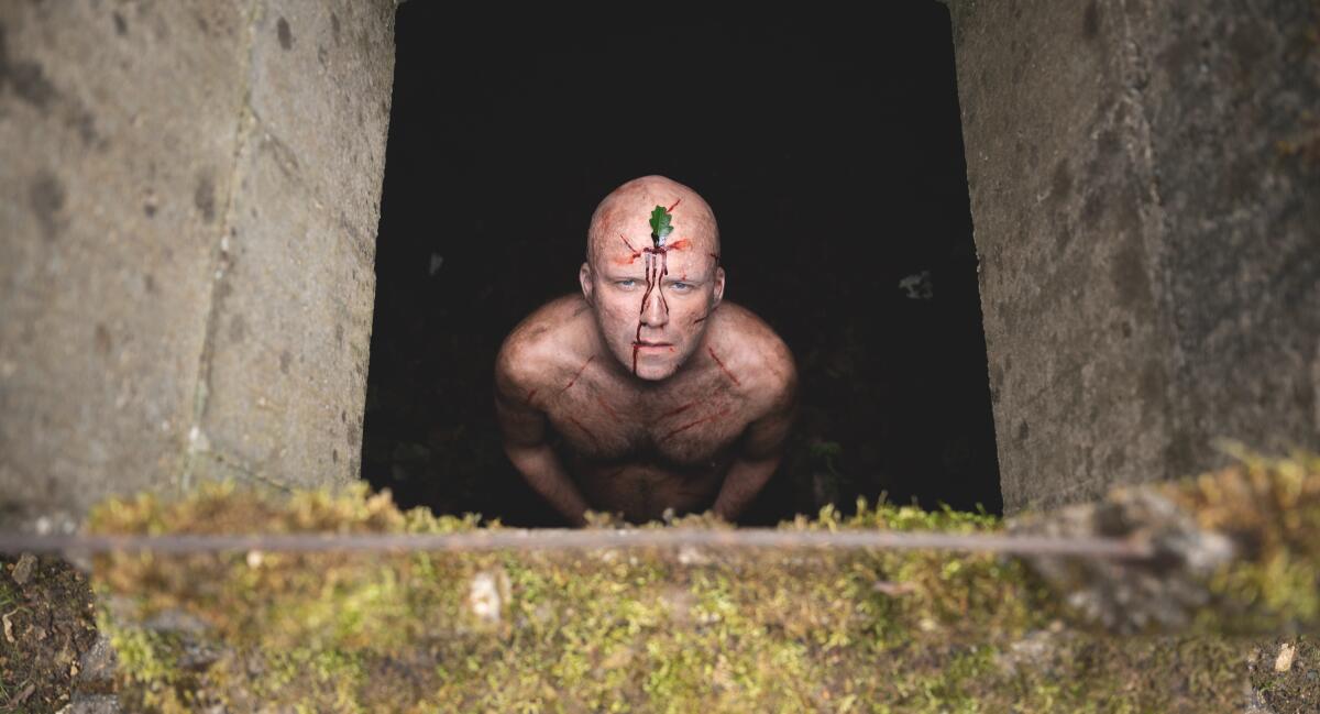 A shirtless man with blood running down his face looks up from a hole in the ground 