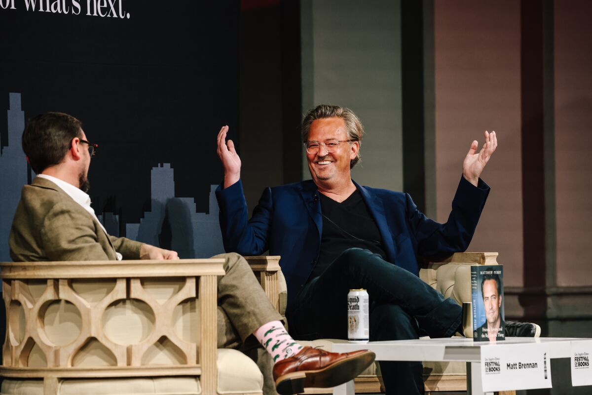 Gülümseyen Matthew Perry, sahnede oturmuş bir röportajcıyla konuşurken kollarını havaya kaldırıyor.