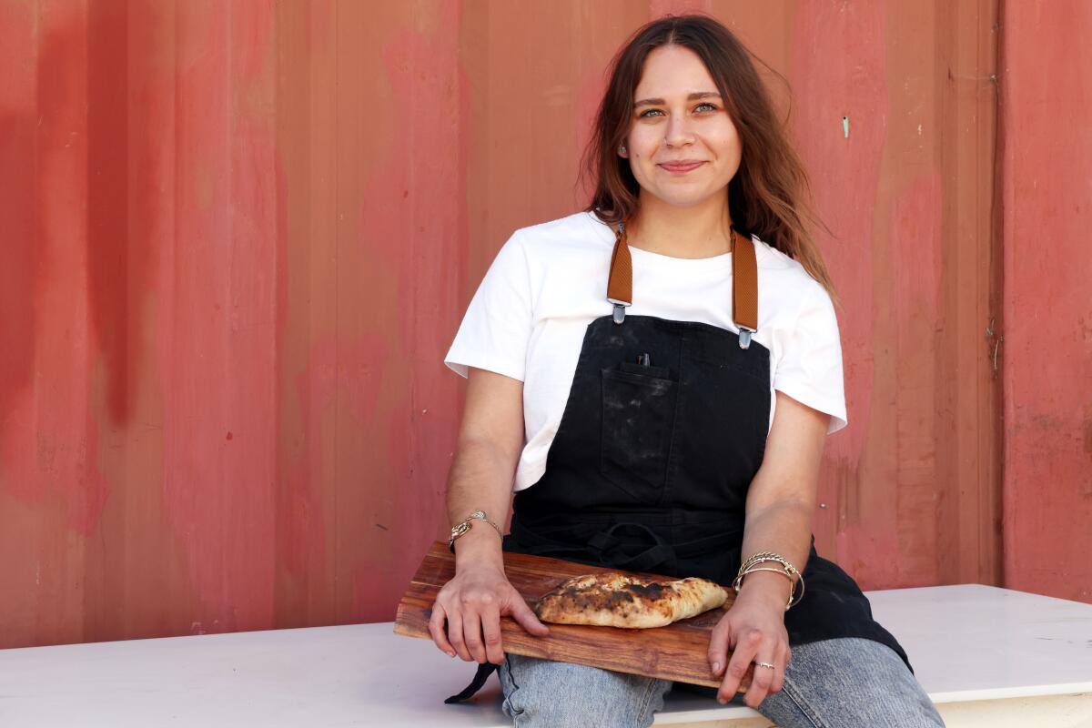 A female chef in an apron sits; in her lap is a wooden board holding one of her khachapuri
