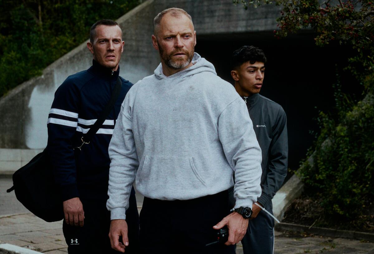 Three men, one in zip-tie handcuffs, peer around.