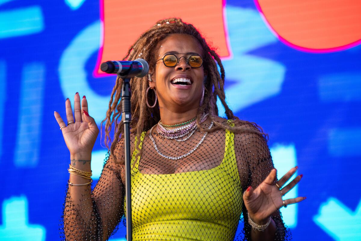 Kelis with braids and sunglasses wearing a neon yellow tank top and a mesh dress while performing on stage