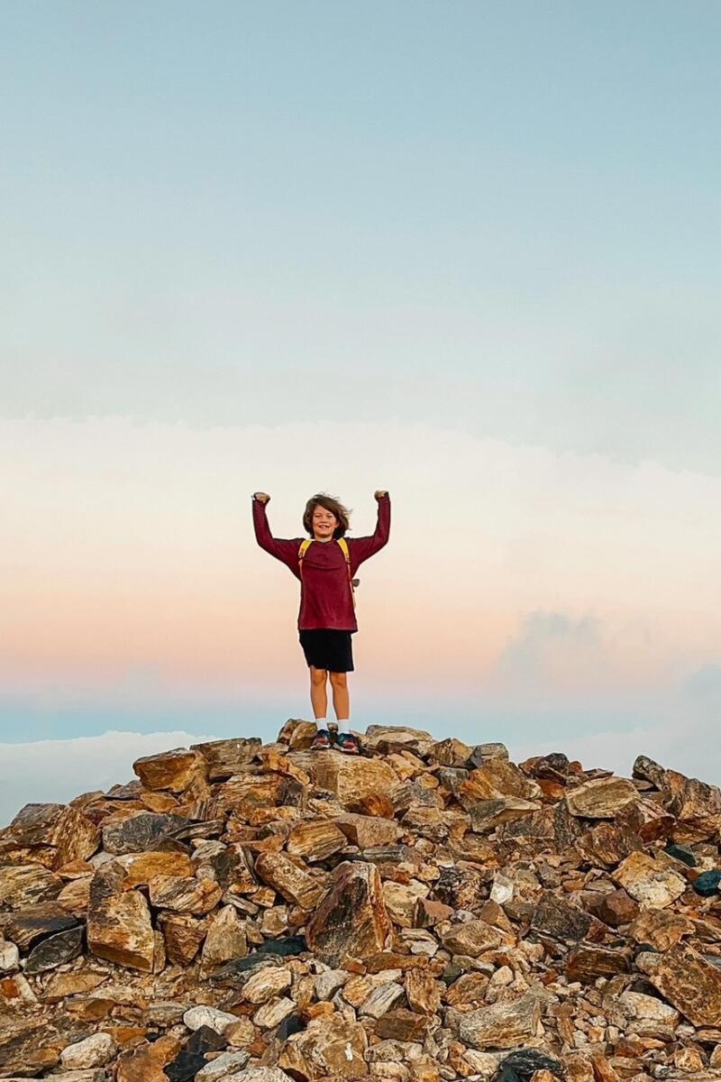 A boy stands at the top of a hill of rocks, his arms raised triumphantly.