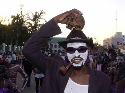 A Haitian man in a costume of the 'God of death' holds a skull to his head during Carnival festivities.